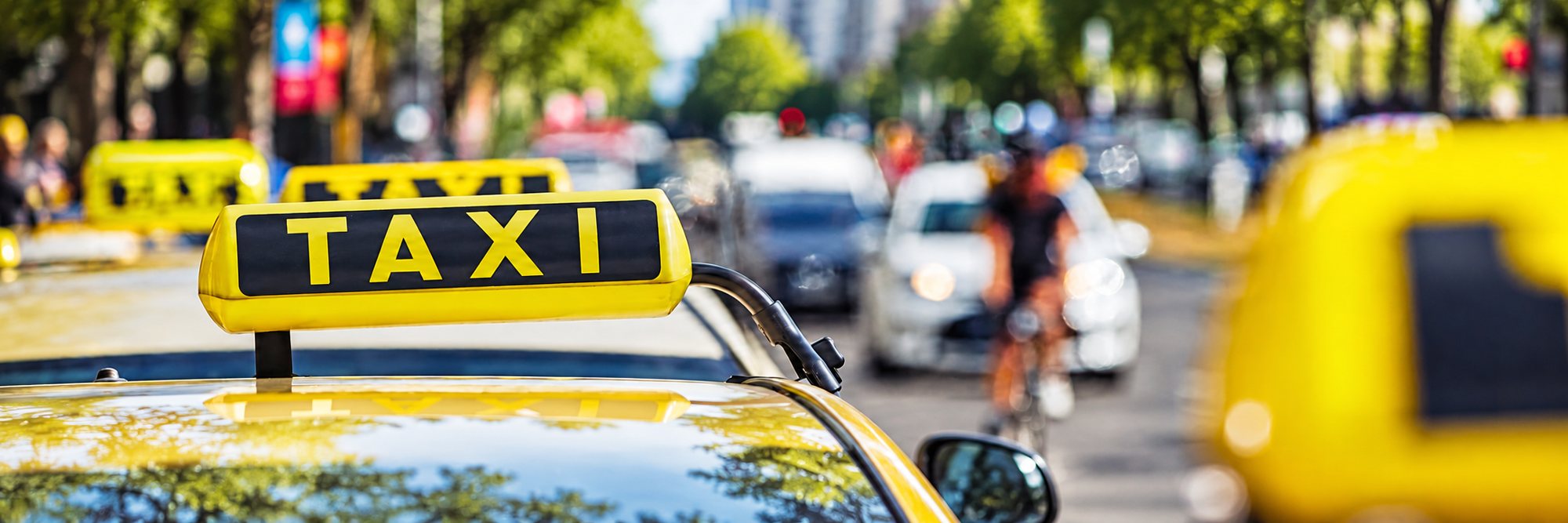 Gros plan sur l'enseigne jaune TAXI posée sur le toit d'un véhicule, avec d'autres taxis et un cycliste flous à l'arrière-plan dans la rue.