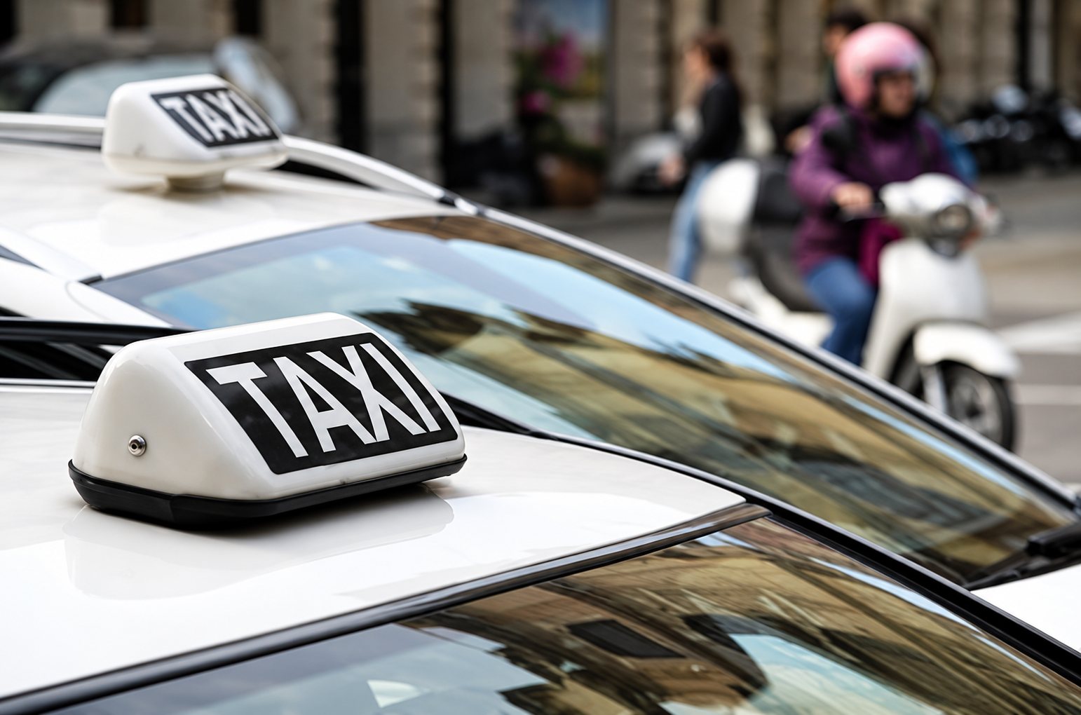 Gros plan sur l'enseigne TAXI posée sur le toit d'un taxi blanc, avec un autre taxi à l'arrière-plan.