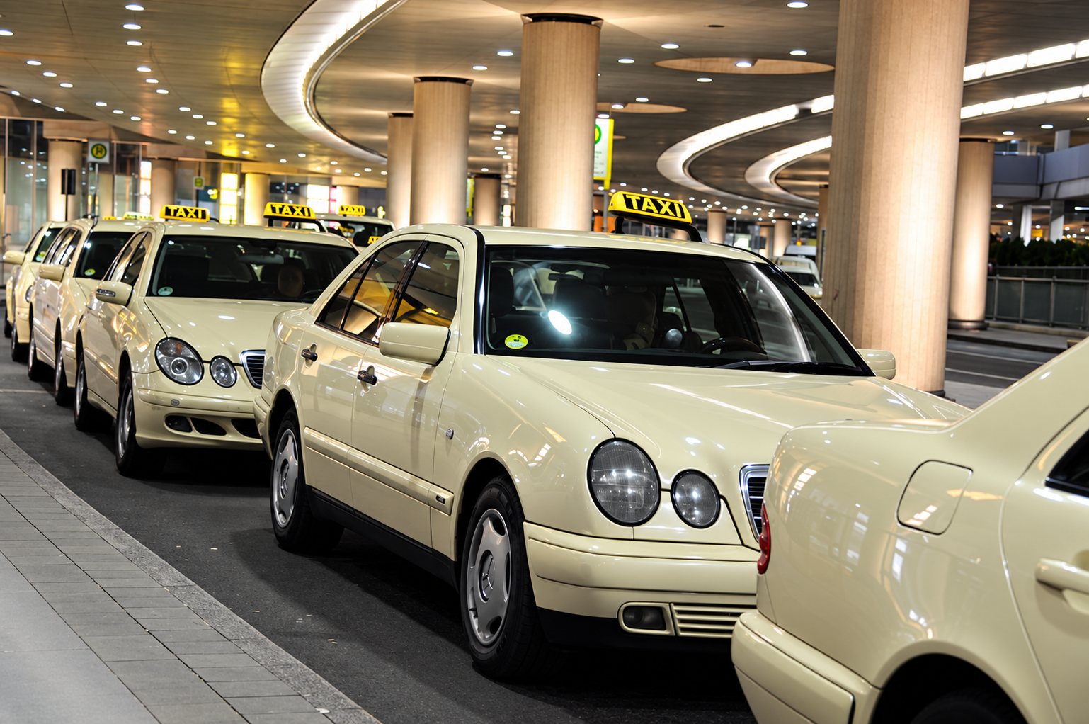 Une file de taxis de couleur crème, avec enseignes lumineuses sur le toit, attend dans une station couverte.