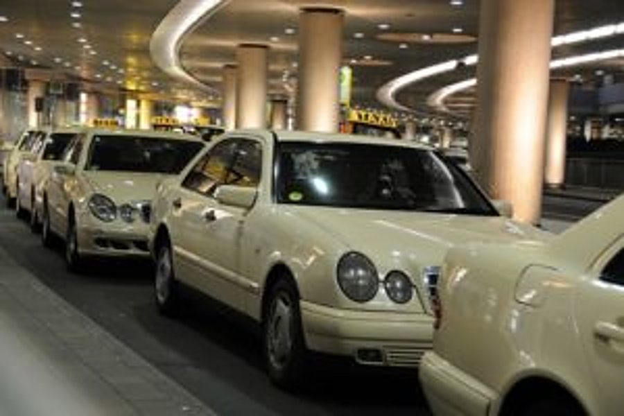 Plusieurs taxis de couleur crème alignés à une station sous un grand auvent, éclairés en soirée.