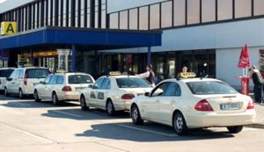 Une file de taxis blancs de type berline est garée devant un bâtiment à auvent bleu.