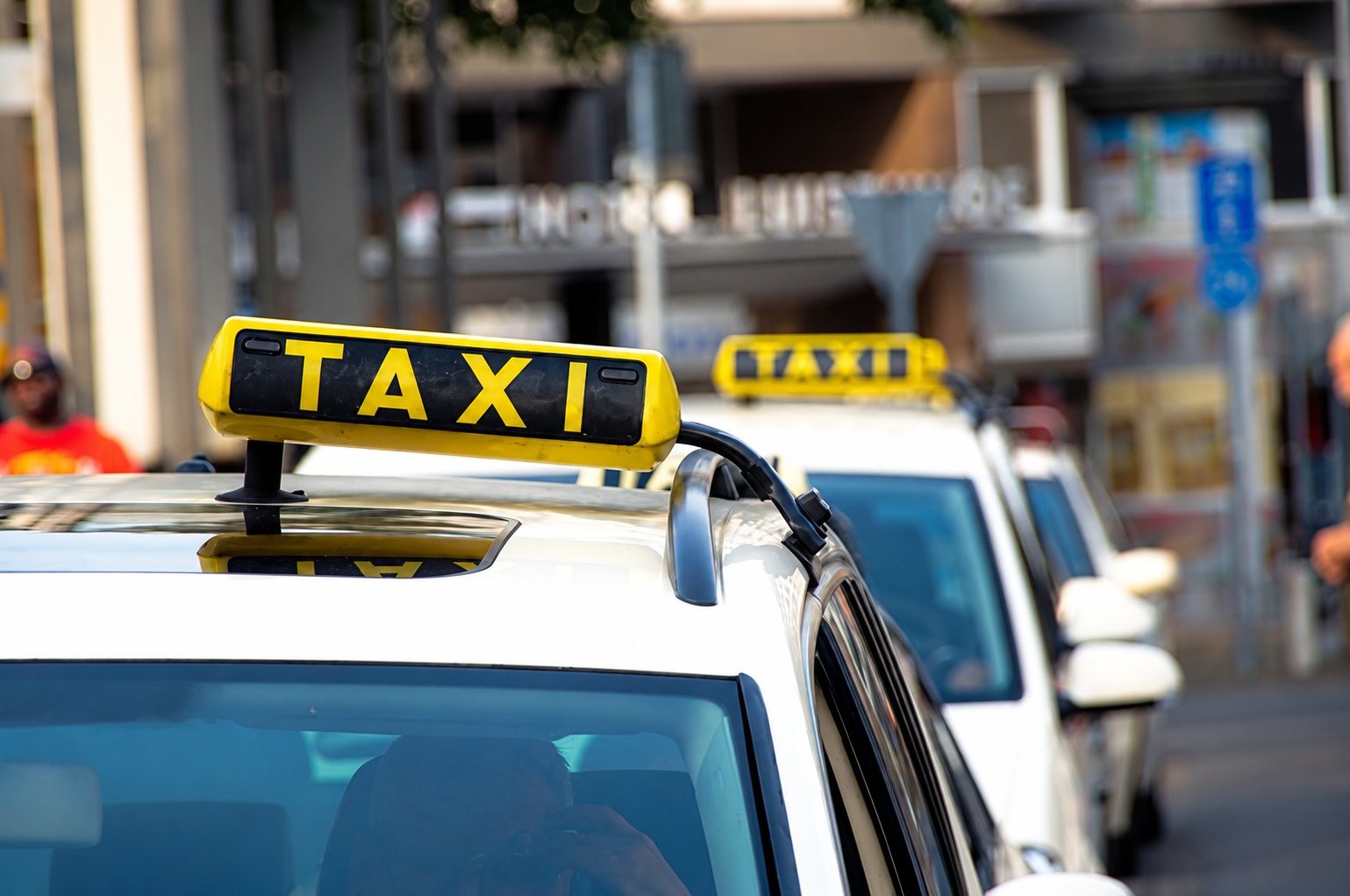 Gros plan sur l'enseigne jaune TAXI posée sur le toit d'une voiture blanche, avec d'autres taxis garés floutés à l'arrière-plan.